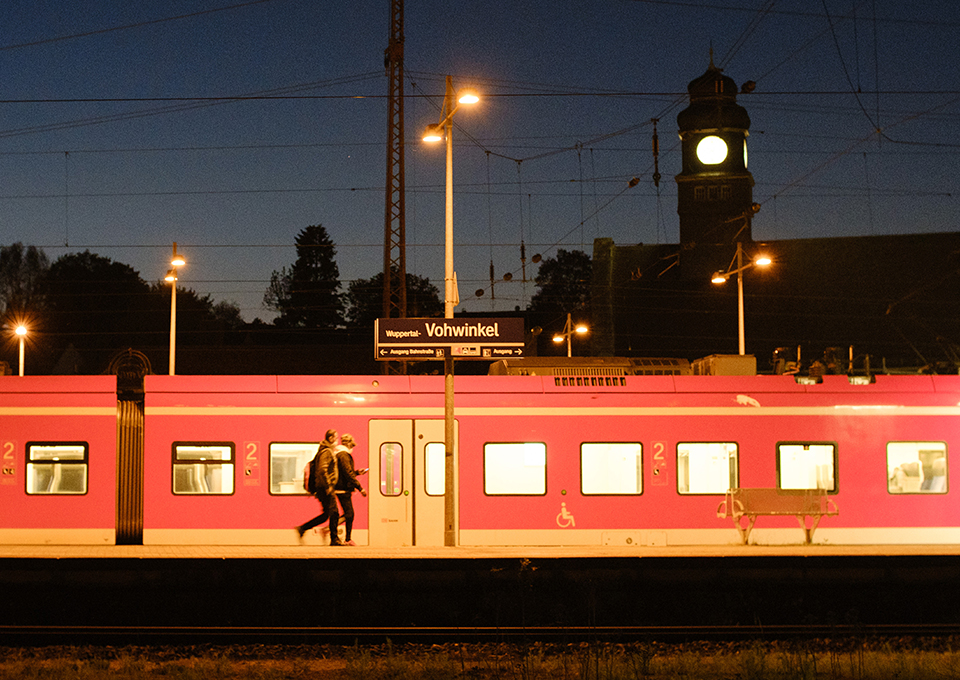 Zug steht in der Dunkelheit an einem beleuchteten Bahnsteig, zwei Fahrgäste laufen auf eine Tür zu