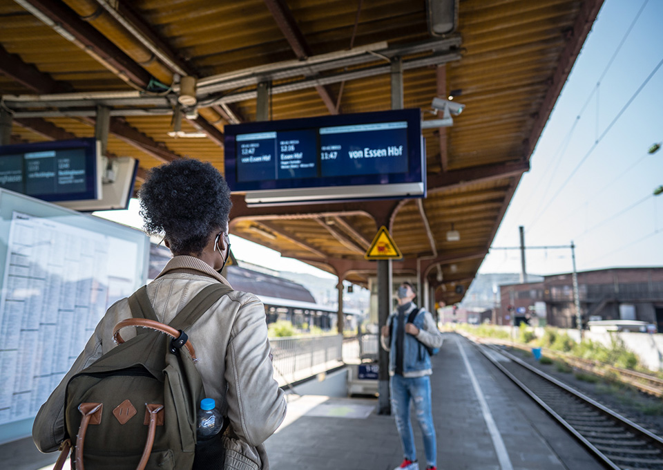 Eine junge Frau mit Rucksack steht am Bahnsteig und blickt auf eine Fahrgastinformationsanzeige. Im Hintergrund ist ein weiterer Fahrgast zu sehen.