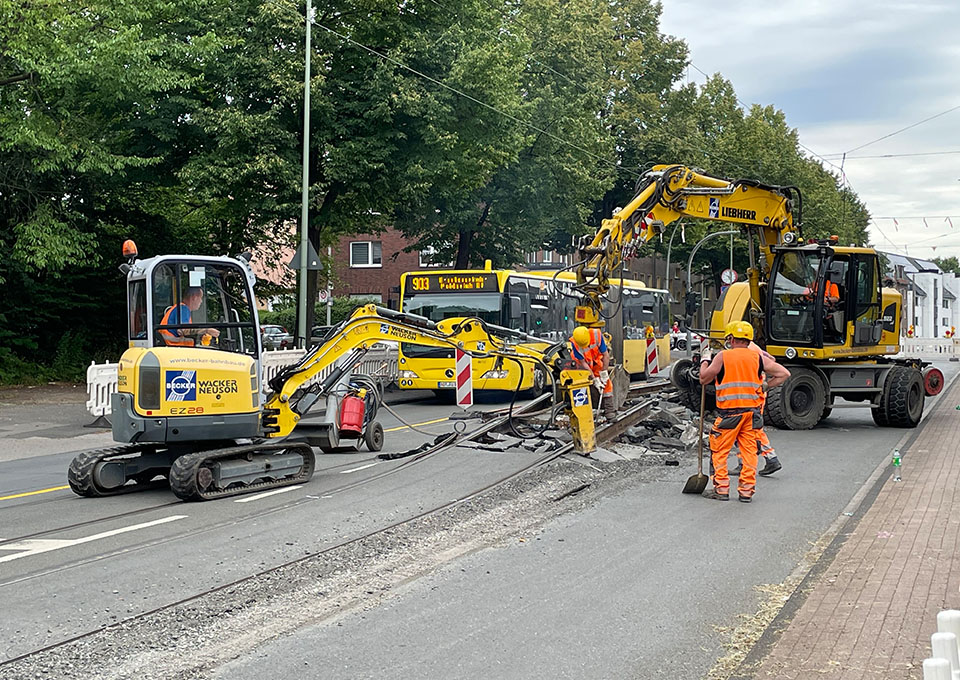 Neubau Haltestelle Brückelstraße: zwei Baumaschinen brechen den Asphalt auf und heben alte Gleise an, Bauarbeiter sind zu sehen, im Hintergrund ein Bus der DVG