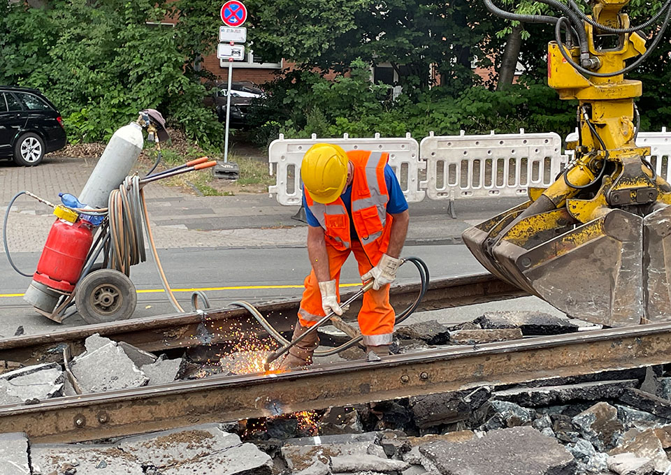Neubau Haltestelle Brückelstraße: Eine Baumaschine hebt alte Gleise an, ein Bauarbeiter verrichtet Schweißarbeiten