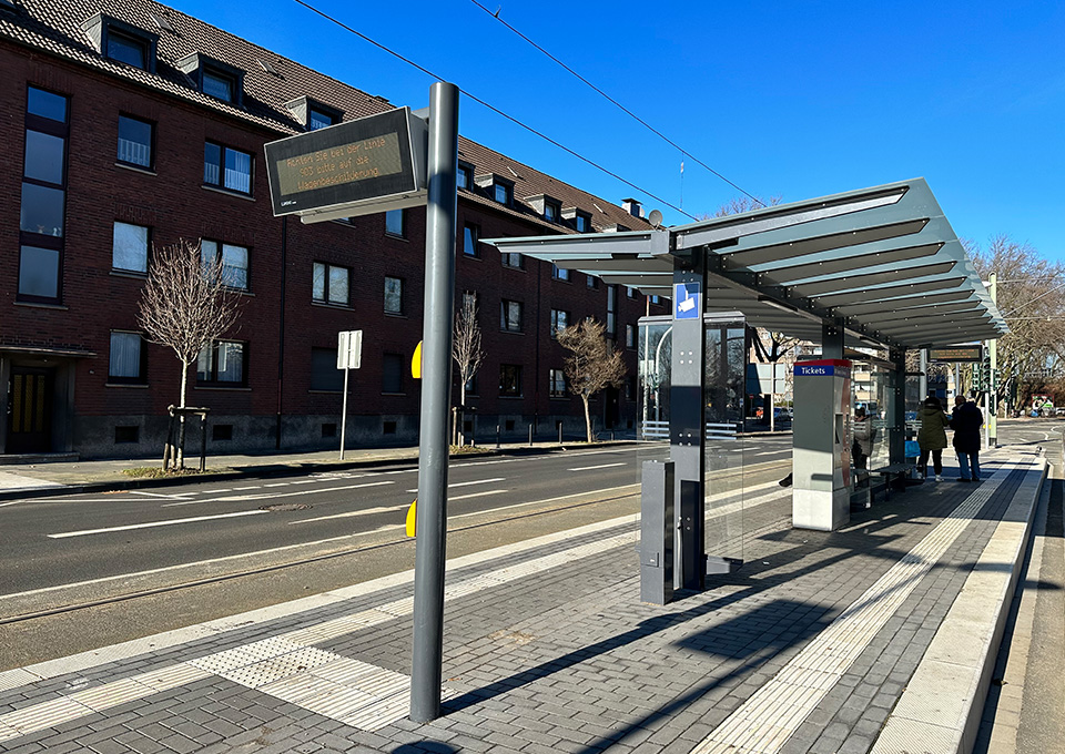 Neubau Haltestelle Brückelstraße: Blick auf die fertige Haltestelle mit Wetterschutz, dynamischen Fahrgastinformation, Ticketautomat und Sitzgelegenheiten, im Hintergrund sind zwei Fahrgäste zu sehen