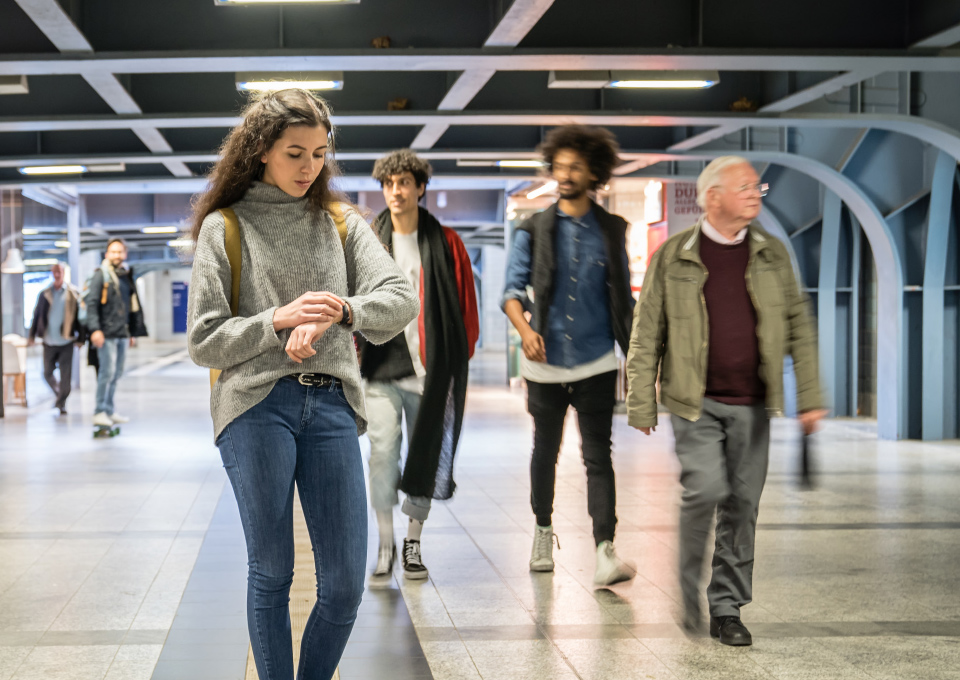 Personen in einem Bahnhof. Eine Frau im Vordergrund schaut auf ihre Armbanduhr