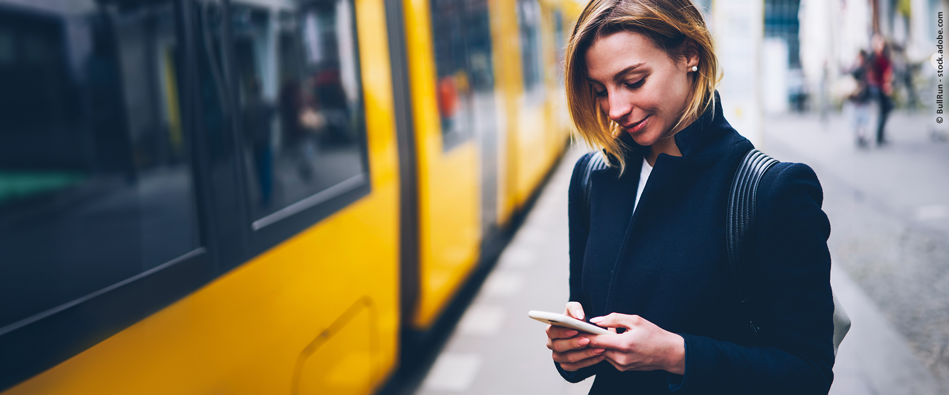 Eine Frau steht an einem Stadtbahn-Bahnsteig und schaut auf ihr Smartphone