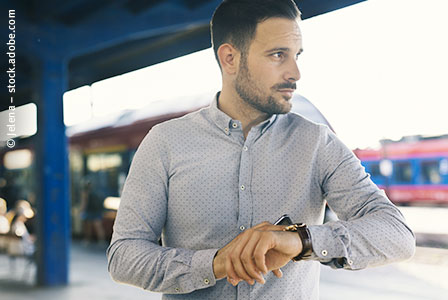 Ein Mann steht an einem Bahnhof und schaut auf seine Uhr.